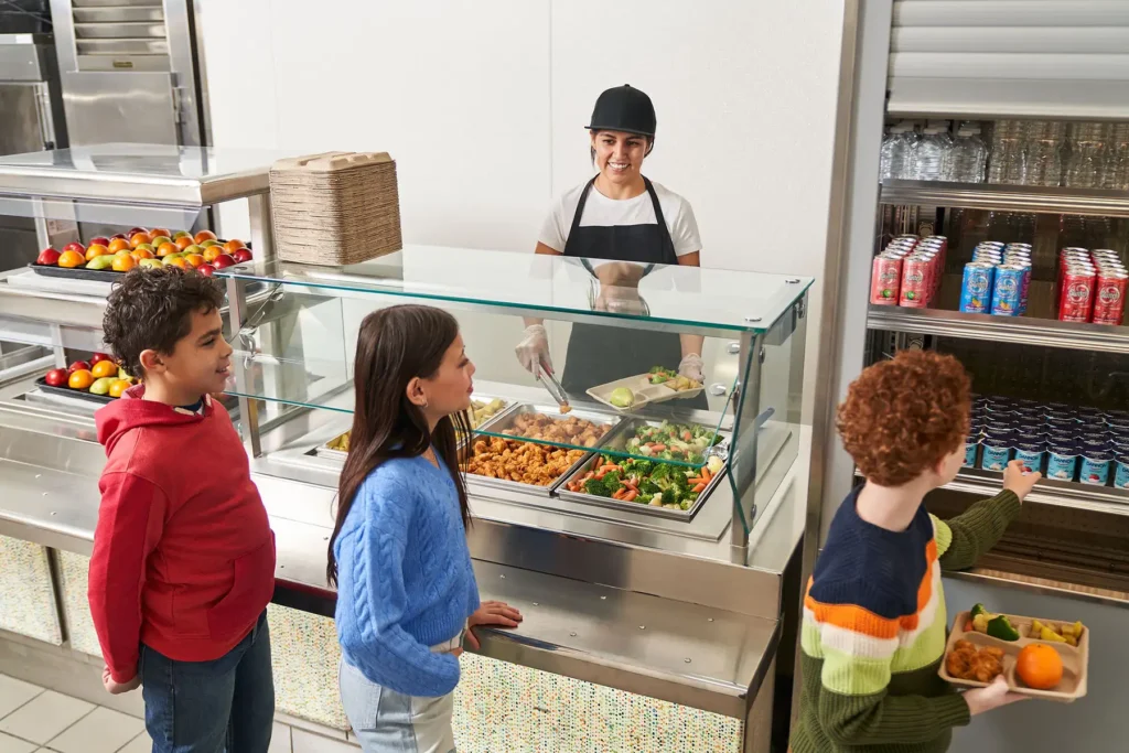 A woman serves hot lunch to young students in a cafeteria