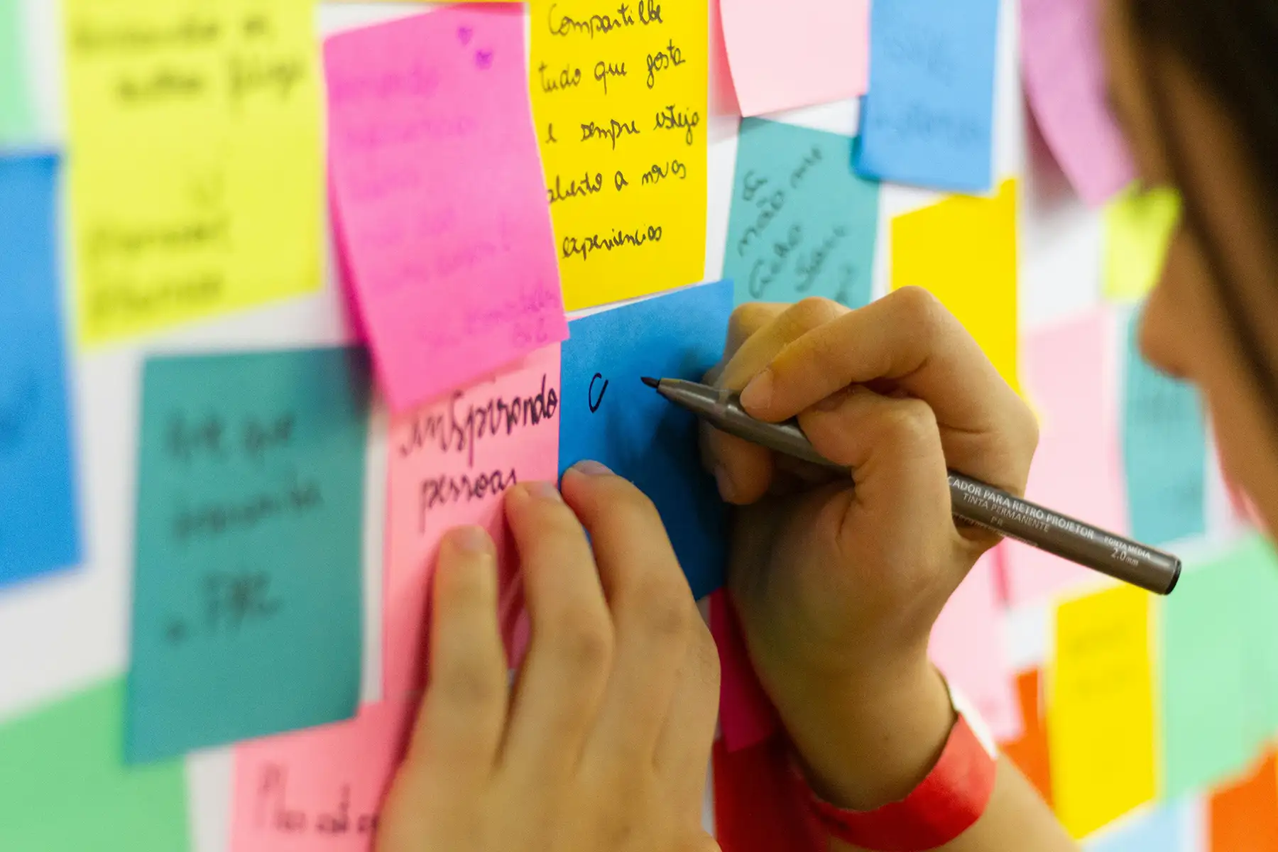 Someone writes on a sticky note on a wall full of other sticky notes with writing on them