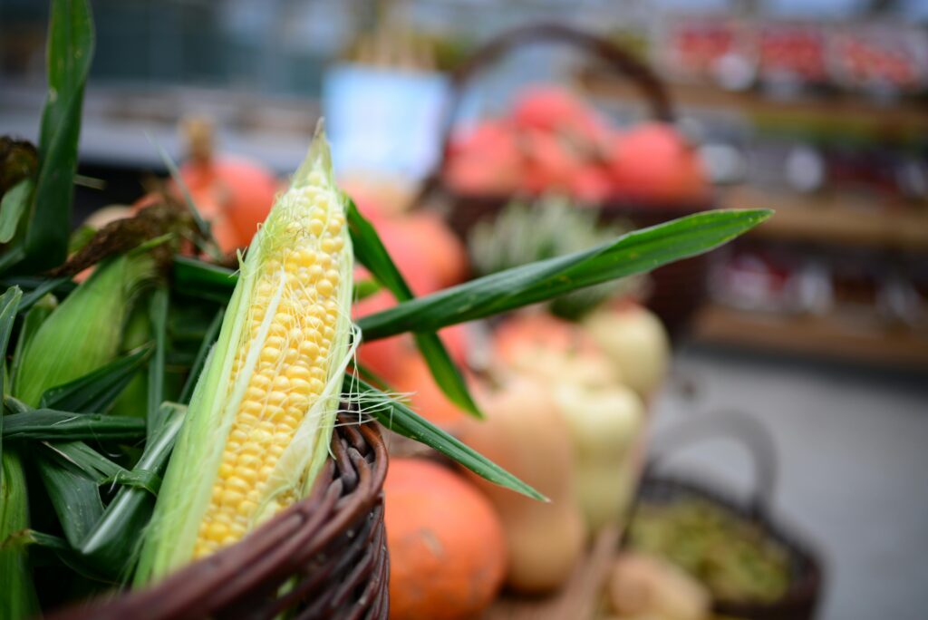 Photo of a basket of corn and other food with fresh produce and other items in a grocery store blurry in the background